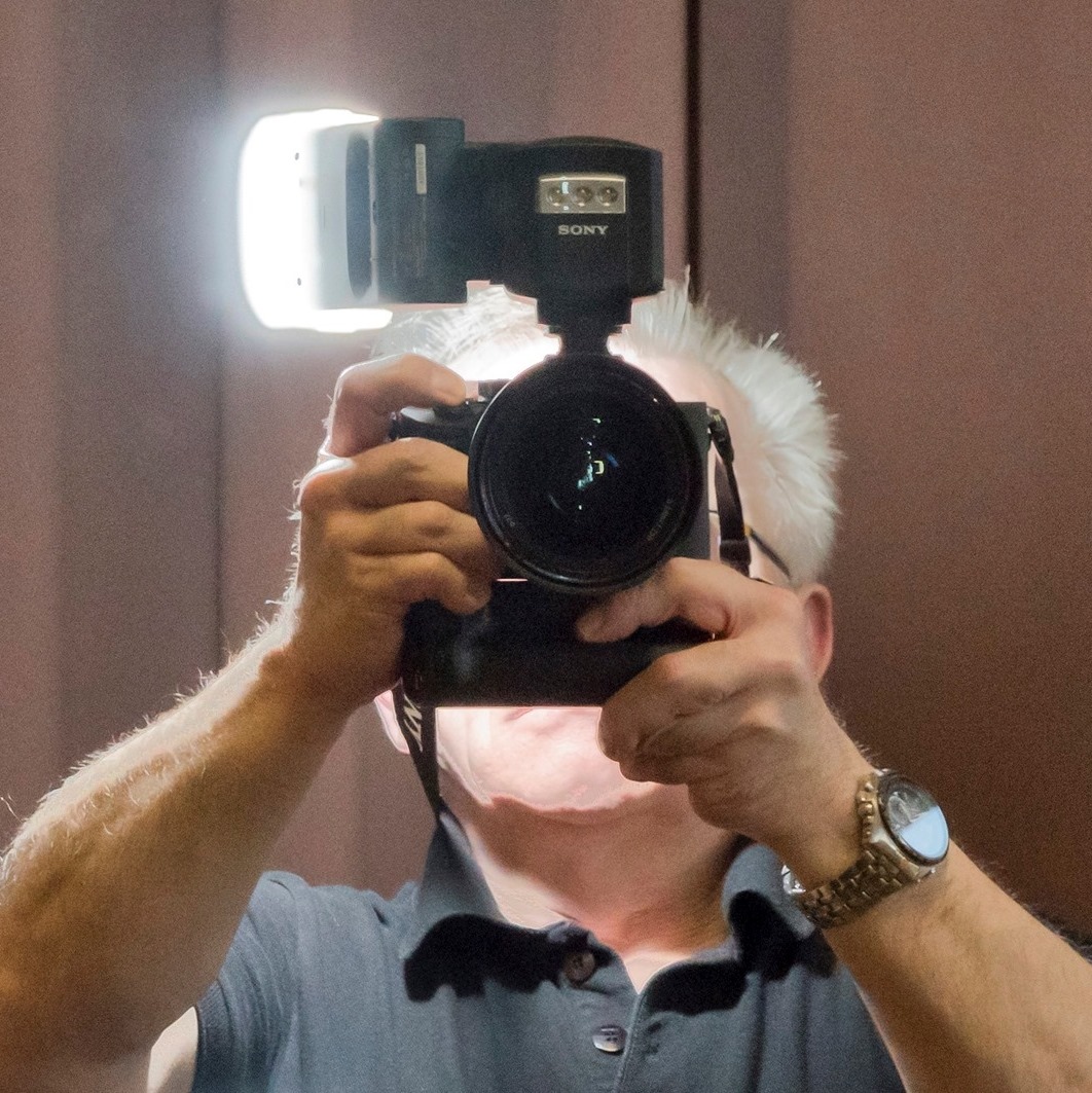 Mężczyzna z siwymi włosami fotografuje siebie w lustrze, trzymając profesjonalny aparat Sony z zamontowaną lampą błyskową, która świeci intensywnym światłem.